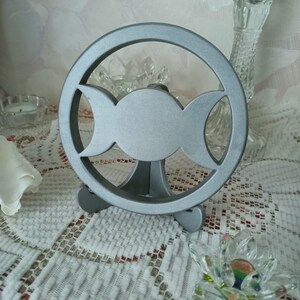 May include: A circular silver-colored metal stand featuring a cutout triple moon symbol. The object consists of a central full circle flanked by two crescent shapes, all enclosed within a larger outer ring, supported by a small tripod base.