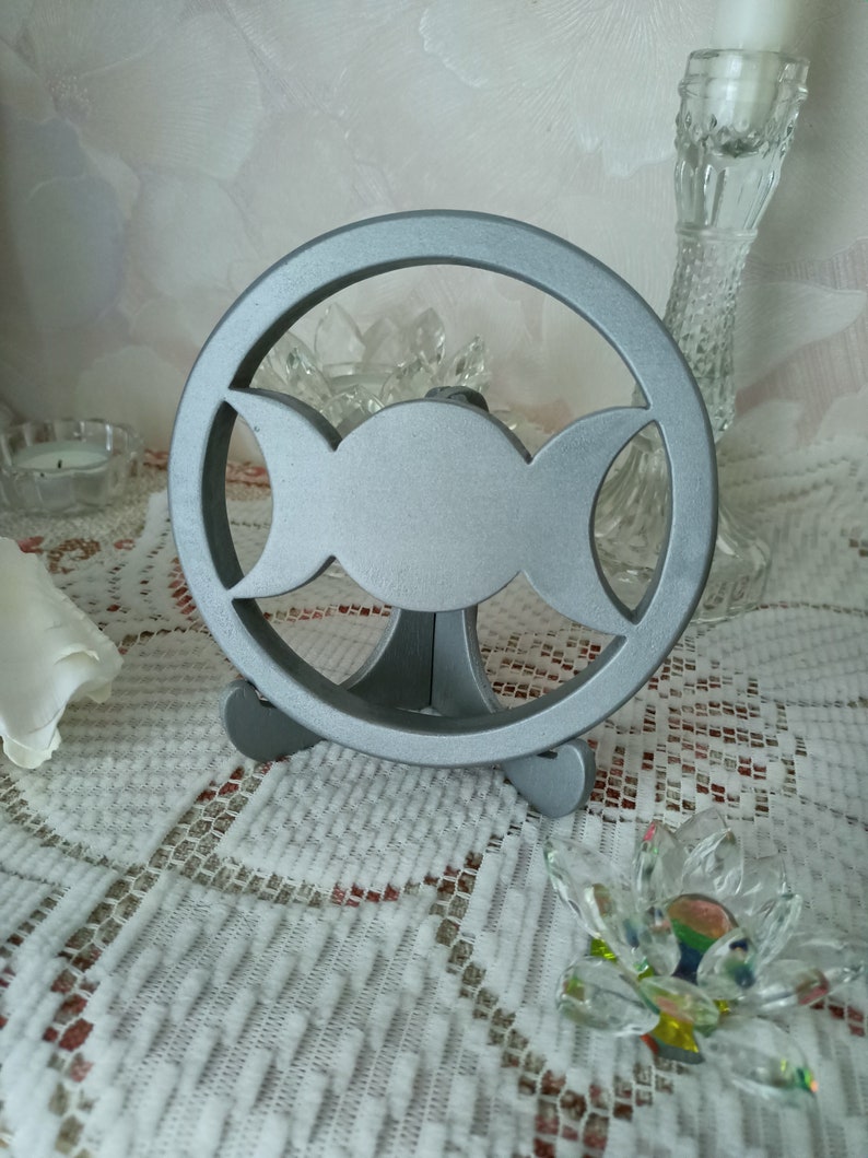 May include: A circular silver-colored metal stand featuring a cutout triple moon symbol. The object consists of a central full circle flanked by two crescent shapes, all enclosed within a larger outer ring, supported by a small tripod base.