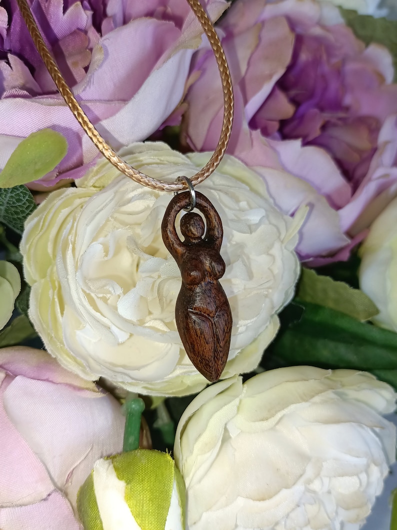 May include: A wooden pendant necklace featuring a carved figure of a woman with arms raised. The pendant is suspended from a tan cord. The background is a soft focus of white and purple flowers.
