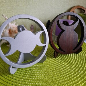 May include: Two circular decorative stands, one finished in a matte silver color and the other in a dark brown wood tone. Each stand features a cutout design in the center, with the silver stand displaying a triple moon symbol and the brown stand showing a stylized owl silhouette, both supported by small integrated feet.