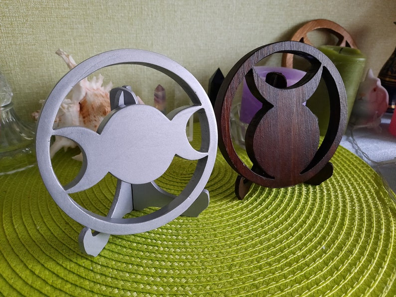 May include: Two circular decorative stands, one finished in a matte silver color and the other in a dark brown wood tone. Each stand features a cutout design in the center, with the silver stand displaying a triple moon symbol and the brown stand showing a stylized owl silhouette, both supported by small integrated feet.