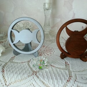 May include: Two circular stands featuring a triple moon symbol cutout. One stand is finished in a matte silver color, while the other is a dark brown wood tone. Each circular frame is supported by a small, integrated base for upright display.