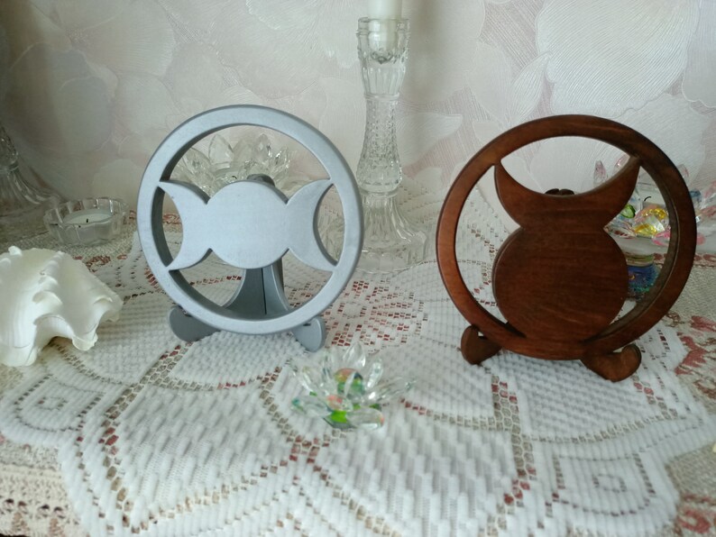 May include: Two circular stands featuring a triple moon symbol cutout. One stand is finished in a matte silver color, while the other is a dark brown wood tone. Each circular frame is supported by a small, integrated base for upright display.