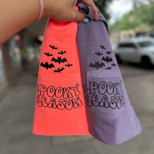 May include: Two t-shirts, one neon orange and one muted purple, each featuring a chest pocket. Both shirts display a black embroidered design of a flock of bats above the pocket and the text "SPOOKY SEASON" in a bold, wavy font on the pocket.