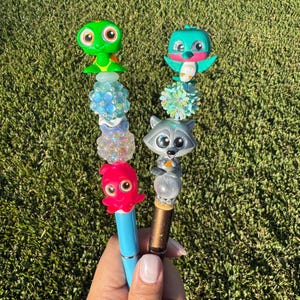 May include: Two decorative pens with colorful animal toppers. One pen features a green turtle, blue and clear beads, and a pink octopus. The other pen has a teal bird, a floral bead, and a gray raccoon.