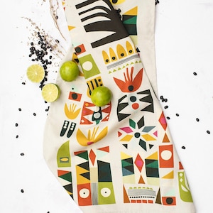 May include: A white kitchen towel with a colourful geometric pattern. The pattern features abstract shapes in shades of red, yellow, green, blue, and black. The towel is folded and lying on a white surface.