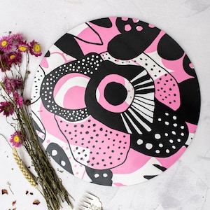 Op de afbeelding: Een rond roze en zwart gepatroneerd bord met een witte achtergrond. Het patroon is abstract en heeft cirkels, stippen en lijnen.