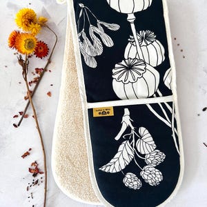 Könnte beinhalten: Schwarzer und weißer Ofenhandschuh mit botanischem Design. Der Handschuh hat einen schwarzen Hintergrund mit weißen Illustrationen von Samenkapseln und Blättern. Die Innenseite ist hellbeige, und der Handschuh hat ein Etikett mit der Aufschrift "Softer + Wild".