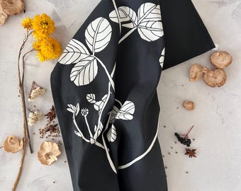 Può includere: Canovaccio da cucina nero con un motivo a foglie botaniche bianche. Il canovaccio è piegato, mostrando il motivo. L'immagine include anche fiori secchi, funghi e spezie, suggerendo un tema ispirato alla natura.