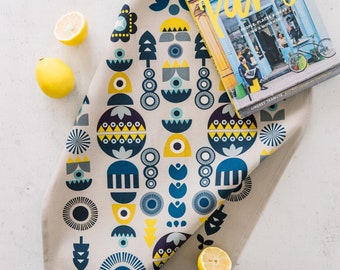 Puede incluir: Un paño de cocina beige con un patrón geométrico en azul, amarillo y gris, junto a un libro titulado "The New Paris" y limones frescos en un cuenco negro. El diseño del paño presenta formas estilizadas.