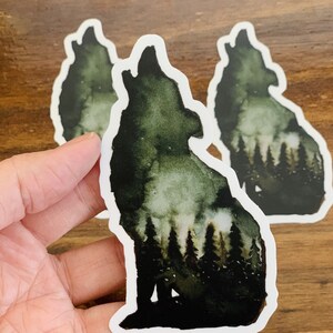 May include: Two watercolour stickers of a wolf howling at the moon with a forest silhouette behind it. The wolf is a dark green colour with a white outline. The stickers are on a wooden surface.