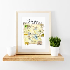 May include: A watercolor illustration of Yellowstone National Park, featuring landmarks like Mammoth Hot Springs, Old Faithful Geyser, and Yellowstone Lake. The illustration is framed in a light wood frame and displayed on a shelf with a potted plant.