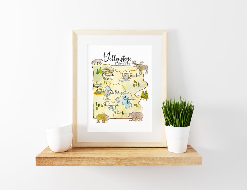 May include: A watercolor illustration of Yellowstone National Park, featuring landmarks like Mammoth Hot Springs, Old Faithful Geyser, and Yellowstone Lake. The illustration is framed in a light wood frame and displayed on a shelf with a potted plant.