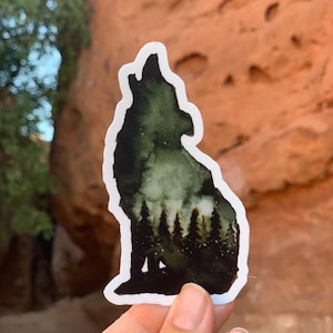 May include: A sticker featuring a silhouette of a wolf howling at the moon. The wolf is outlined in white and filled with a dark green watercolour design. The background of the sticker is a dark forest scene with trees and a starry sky.