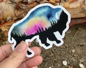 May include: A sticker featuring a bison silhouette with a colorful watercolor design. The bison is black, with a vibrant sky of blue, pink, and yellow. The design includes a forest silhouette and a crescent moon.