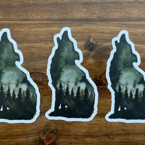 May include: Three watercolour stickers of a wolf howling at the moon with a forest silhouette inside the wolf's body. The stickers are on a brown wooden surface.