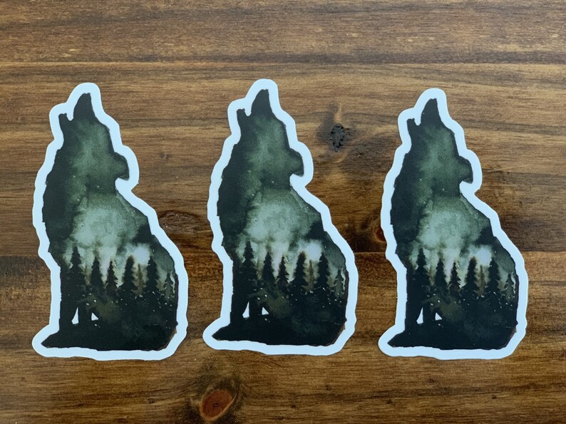May include: Three watercolour stickers of a wolf howling at the moon with a forest silhouette inside the wolf's body. The stickers are on a brown wooden surface.