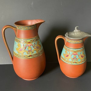 May include: Two terracotta pitchers with a blue and gold design. The larger pitcher has a wide mouth and a handle. The smaller pitcher has a lid made of pewter.
