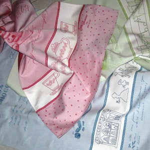 Può includere: Tre asciugamani da cucina con diversi disegni. Il primo asciugamano è rosa con pois bianchi e un disegno ricamato bianco di una tazza da tè. Il secondo asciugamano è azzurro con testo ricamato bianco e un disegno di un negozio di cioccolato. Il terzo asciugamano è verde con testo ricamato bianco e un disegno di alberi.