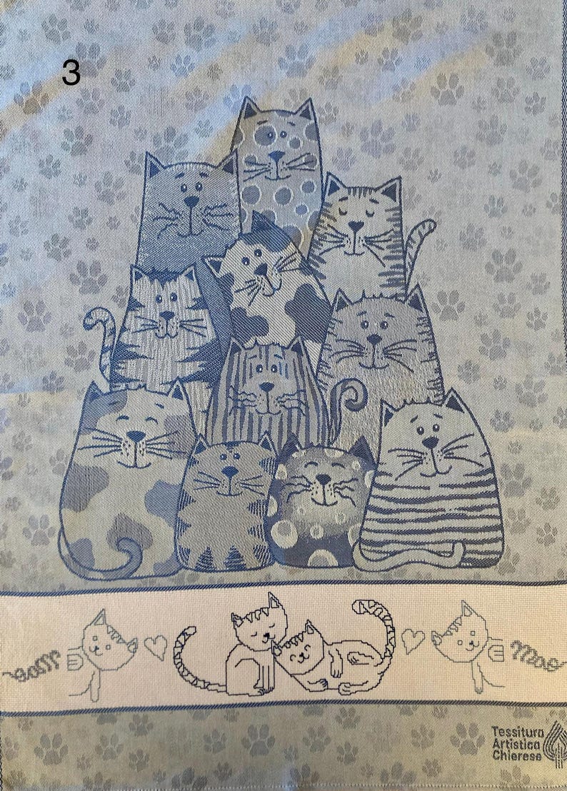 Peut inclure: Une serviette de cuisine bleue et blanche avec un motif de chats. Les chats sont tous de couleurs diff&eacute;rentes et ont des expressions diff&eacute;rentes. La serviette porte le num&eacute;ro 3. Le texte "Tessitura Artistica Chierese" est imprim&eacute; en bas de la serviette.