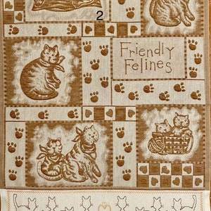 May include: A beige kitchen towel with a patchwork design featuring brown cats and paw prints. The text "Friendly Felines" is printed in the center of the towel. The bottom border of the towel features a row of embroidered cats.