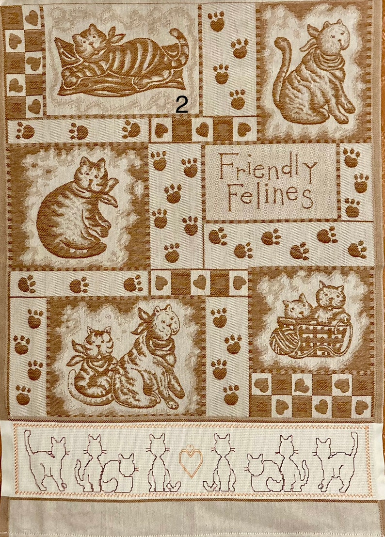 May include: A beige kitchen towel with a patchwork design featuring brown cats and paw prints. The text "Friendly Felines" is printed in the center of the towel. The bottom border of the towel features a row of embroidered cats.