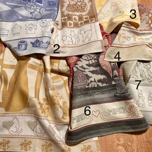 May include: Seven kitchen towels with cat designs. The towels are in various colors, including beige, blue, gray, and white. The designs include cats in a kitchen, cats in a basket, cats with paw prints, and cats with hearts. The towels are all made of a soft, absorbent material.