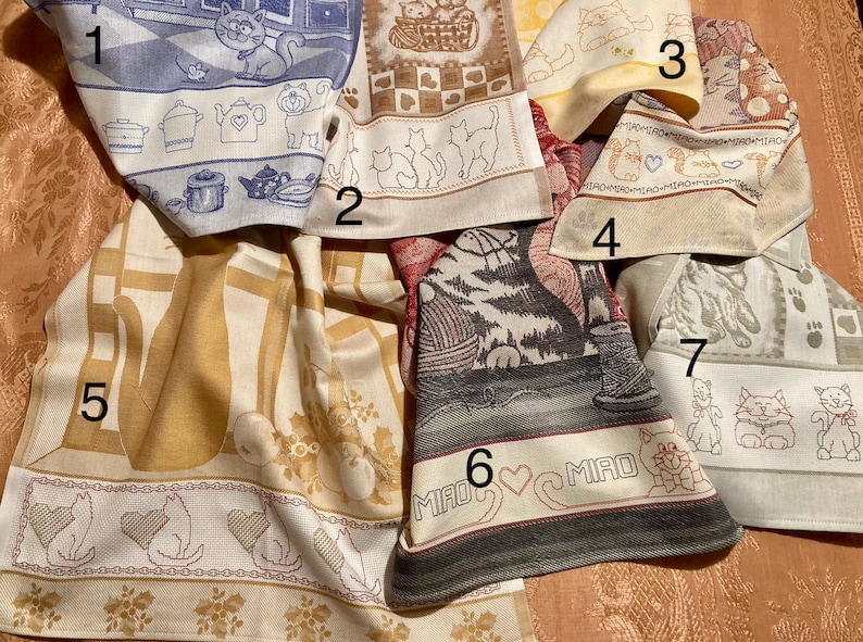 May include: Seven kitchen towels with cat designs. The towels are in various colors, including beige, blue, gray, and white. The designs include cats in a kitchen, cats in a basket, cats with paw prints, and cats with hearts. The towels are all made of a soft, absorbent material.