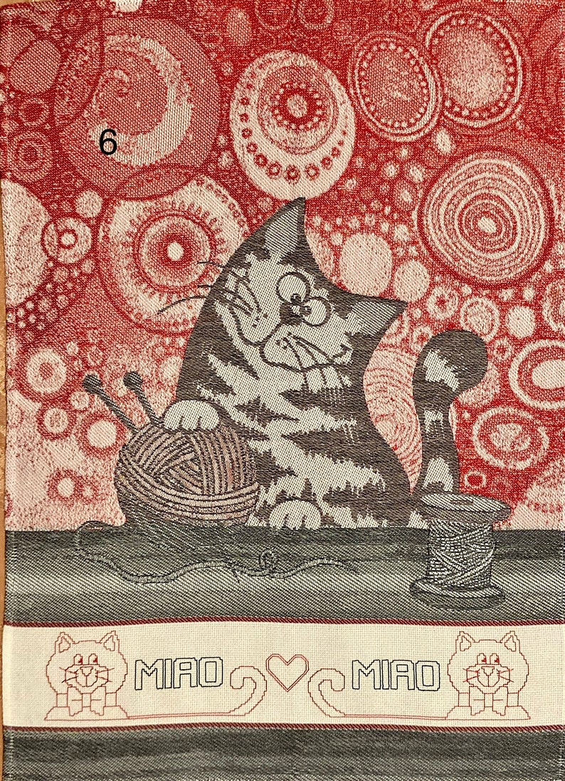 May include: A red and white kitchen towel with a gray cat holding a ball of yarn. The cat is sitting on a table with a spool of thread. The towel has a border with the words "MIAO &hearts; MIAO" and two small cats.