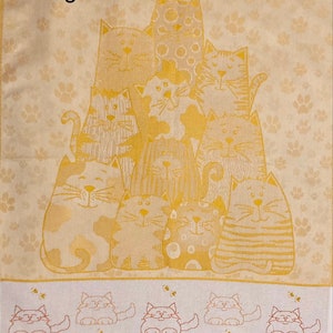May include: A beige kitchen towel with a pattern of paw prints and a group of 10 cats in a gold color. The cats are arranged in a pyramid shape with the largest cat at the top. The towel has a border of smaller cats in a row at the bottom. The text "Tessitura Artistica Chierese" is printed in the bottom right corner.