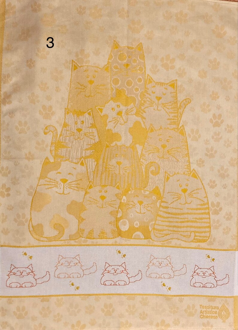 May include: A beige kitchen towel with a pattern of paw prints and a group of 10 cats in a gold color. The cats are arranged in a pyramid shape with the largest cat at the top. The towel has a border of smaller cats in a row at the bottom. The text "Tessitura Artistica Chierese" is printed in the bottom right corner.