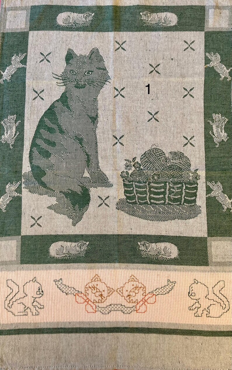 Peut inclure: Une serviette de cuisine verte et blanche avec un chat, un panier de laine et le chiffre "1" brod&eacute; dessus. La serviette a une bordure de carr&eacute;s verts et blancs.