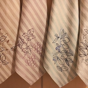 Può includere: Tre asciugamani da cucina con motivi a righe e disegni di frutta ricamati. Gli asciugamani sono beige, azzurro e bianco con ricami neri. I disegni includono uva, melograni e fragole.