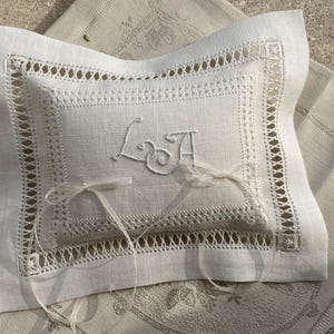 May include: A white linen ring pillow with embroidered initials and a delicate ribbon. The pillow features intricate cutwork and a decorative border. A small bow is tied on the front. The pillow is placed on a patterned linen surface.