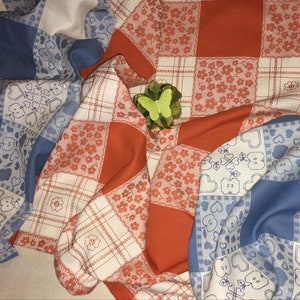 Può includere: Due tovaglie a quadri, una blu e bianca con un motivo a cuore e l'altra arancione e bianca con un motivo floreale. Una farfalla verde è posizionata sopra le tovaglie.