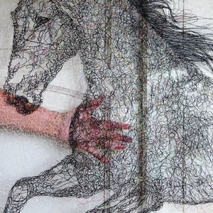 May include: A black horse sculpture made of thread, with a hand reaching out to touch it. The horse is in mid-air, with its legs outstretched. The sculpture is on a white background.