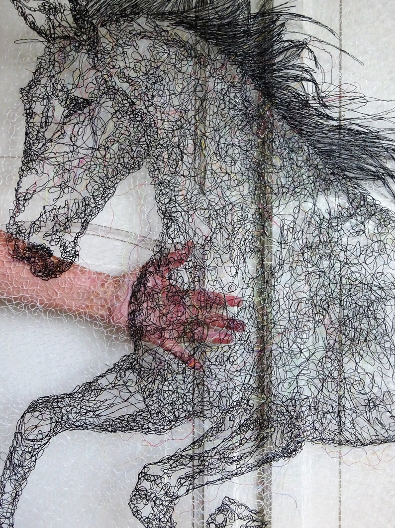 May include: A black horse sculpture made of thread, with a hand reaching out to touch it. The horse is in mid-air, with its legs outstretched. The sculpture is on a white background.