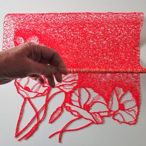 Puede incluir: Una pieza de tejido rojo, intrincada y tejida a crochet con un dise&ntilde;o floral. El tejido est&aacute; estirado en una varilla de madera.
