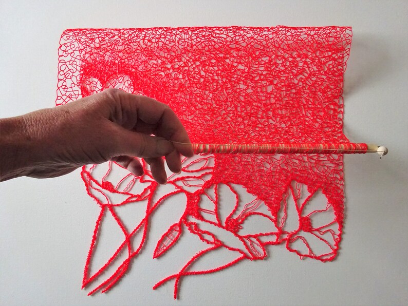 Puede incluir: Una pieza de tejido rojo, intrincada y tejida a crochet con un dise&ntilde;o floral. El tejido est&aacute; estirado en una varilla de madera.