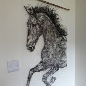 May include: Black and white wire sculpture of a horse's head and front legs, hanging on a white wall. The horse is depicted in mid-air, with its legs extended. The sculpture is made of fine wire, creating a detailed and intricate design.