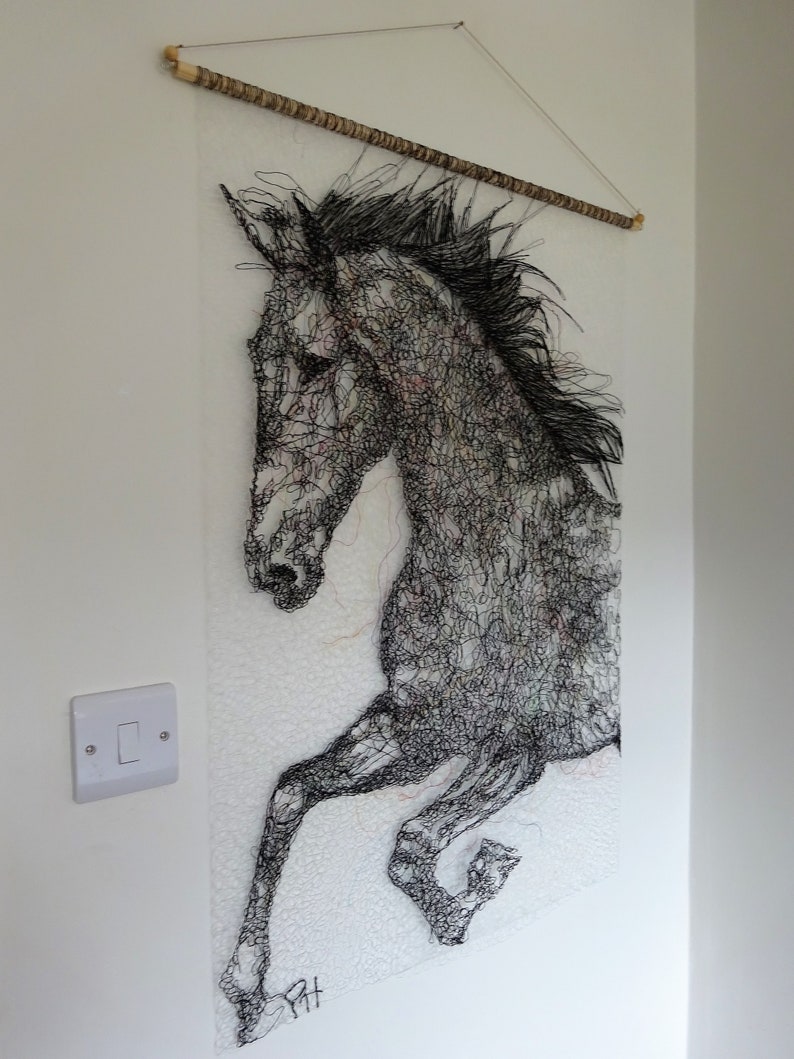 May include: Black and white wire sculpture of a horse's head and front legs, hanging on a white wall. The horse is depicted in mid-air, with its legs extended. The sculpture is made of fine wire, creating a detailed and intricate design.
