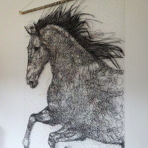 May include: A black and white wire sculpture of a horse in mid-air, with its mane flowing in the wind. The sculpture is mounted on a white wall with a wooden dowel rod at the top.