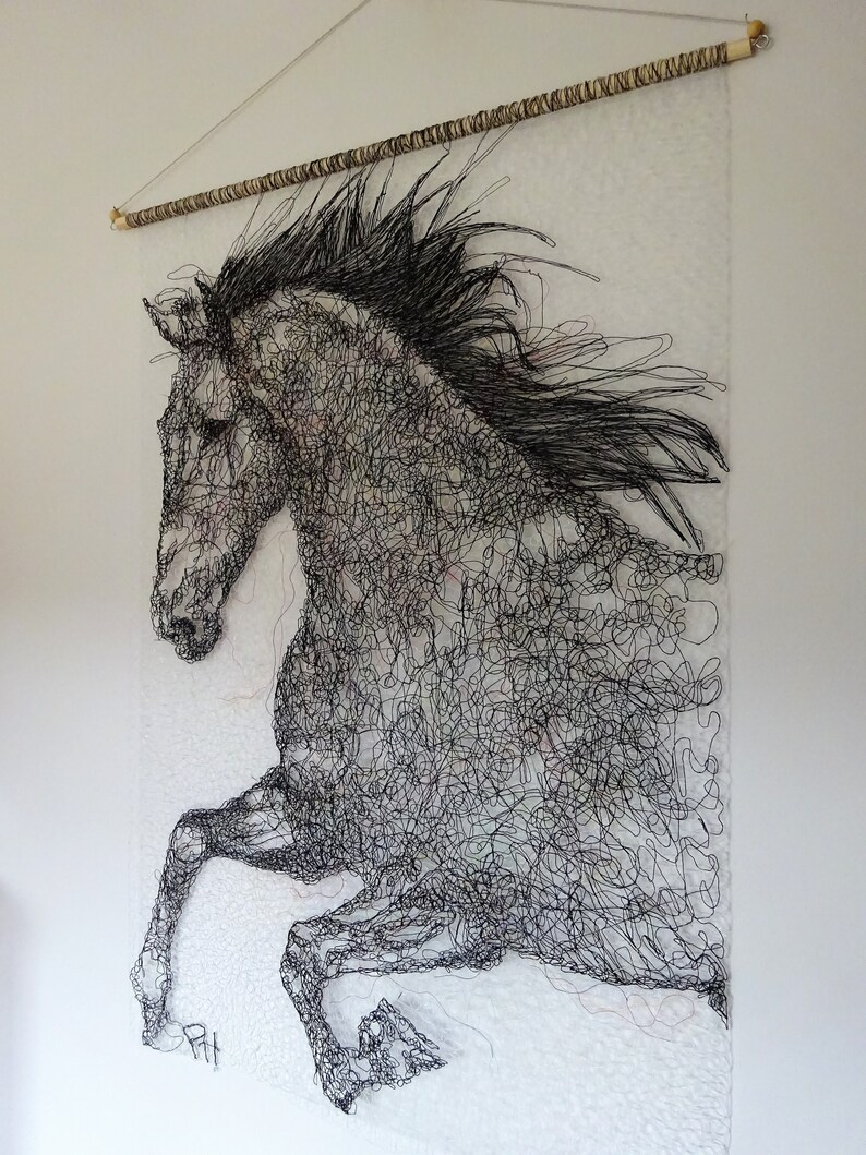 May include: A black and white wire sculpture of a horse in mid-air, with its mane flowing in the wind. The sculpture is mounted on a white wall with a wooden dowel rod at the top.