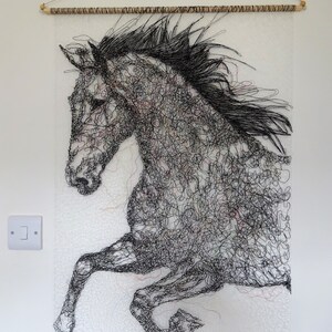 May include: A black and white wire sculpture of a horse in mid-air, with its legs extended. The sculpture is mounted on a white wall with a wooden dowel rod at the top.