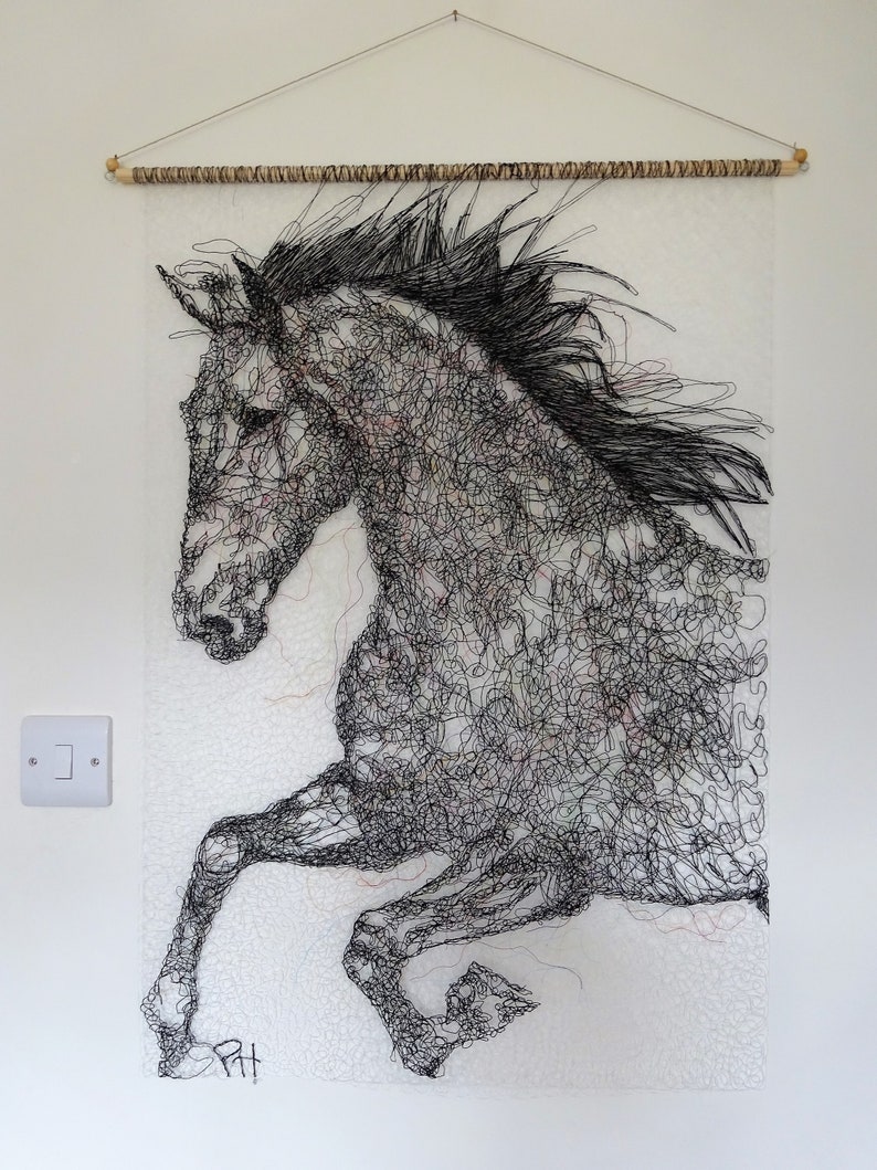 May include: A black and white wire sculpture of a horse in mid-air, with its legs extended. The sculpture is mounted on a white wall with a wooden dowel rod at the top.