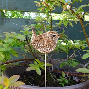 Op de afbeelding: Een decoratieve vogel tuinsteker. De vogel is bruin en wit met een gedetailleerde geweven textuur. Hij is gemonteerd op een houten stok en geplaatst in een potplant met groene bladeren en kleine witte bloemen.