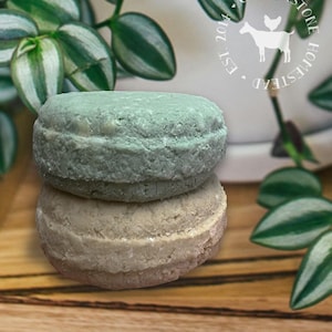 May include: Two round, solid shampoo bars stacked on top of each other. The top bar is light blue and the bottom bar is light brown. The bars are on a wooden surface.
