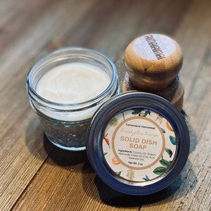 May include: A glass jar with a silver lid containing a white solid dish soap. The lid has a label that reads "Cornerstone Homestead Eucalyptus Orange Solid Dish Soap" and lists the ingredients: Coconut Oil, Sodium Hydroxide, Castor Oil, Citric Acid, Essential Oils. The jar is sitting on a wooden surface next to a wooden scrubber with the word "AIMAIAIMAI" printed on it.