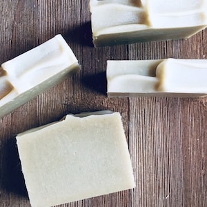 May include: Four bars of handmade soap, each with a light green base and a white swirl design. The soap bars are arranged on a wooden surface.