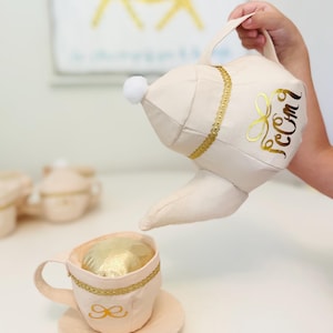 May include: A beige fabric toy teapot pouring into a matching teacup and saucer. The teapot and teacup are trimmed with gold ribbon and a gold bow design. The teapot has a white pom-pom and gold lettering.